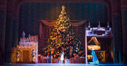 A ballet dancer stands in front of a giant Christmas tree surrounded by presents