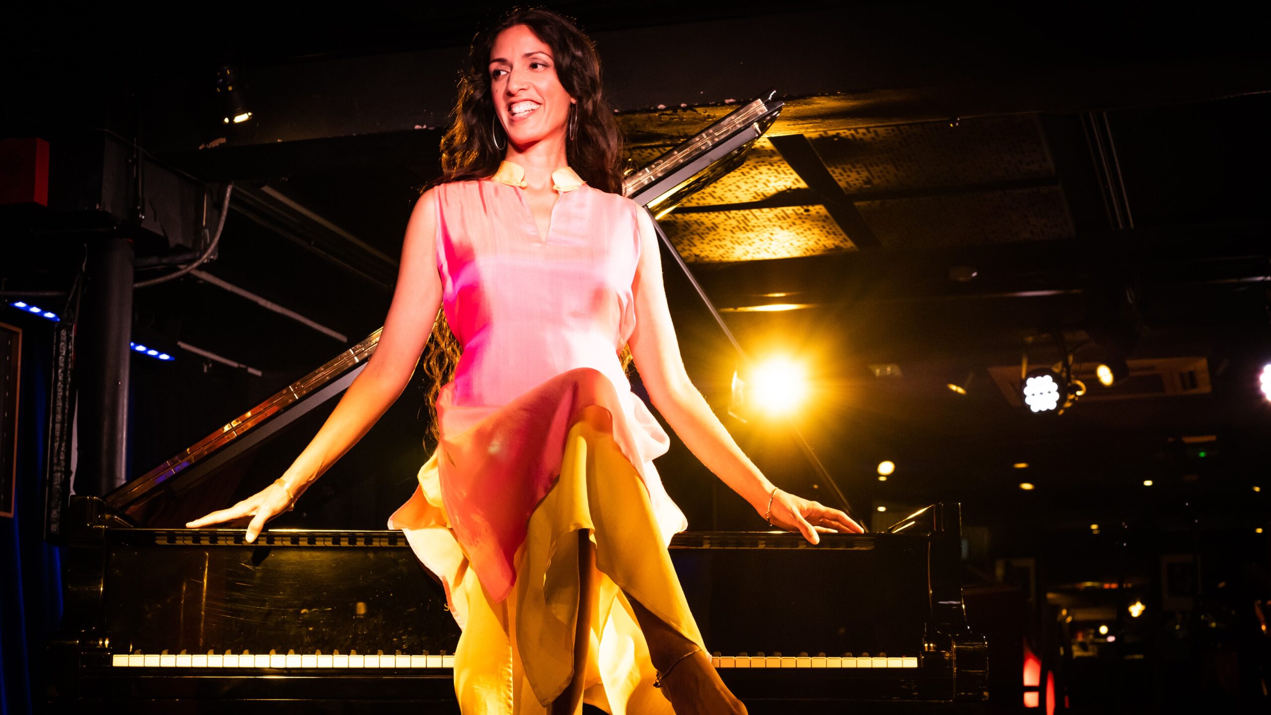 Jazz pianist Zoe Rahmann sits on top of a piano in a flamboyant pink dress, smiling with her head slightly turned to the left