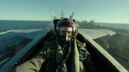 Actor Tom Cruise sits in a fighter jet as the pilot, wearing a blue and red helmet