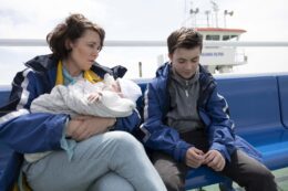 Actress Olivia Colman sits on a bench, holding a baby, next to a young boy. There is a boat and the sea in the background