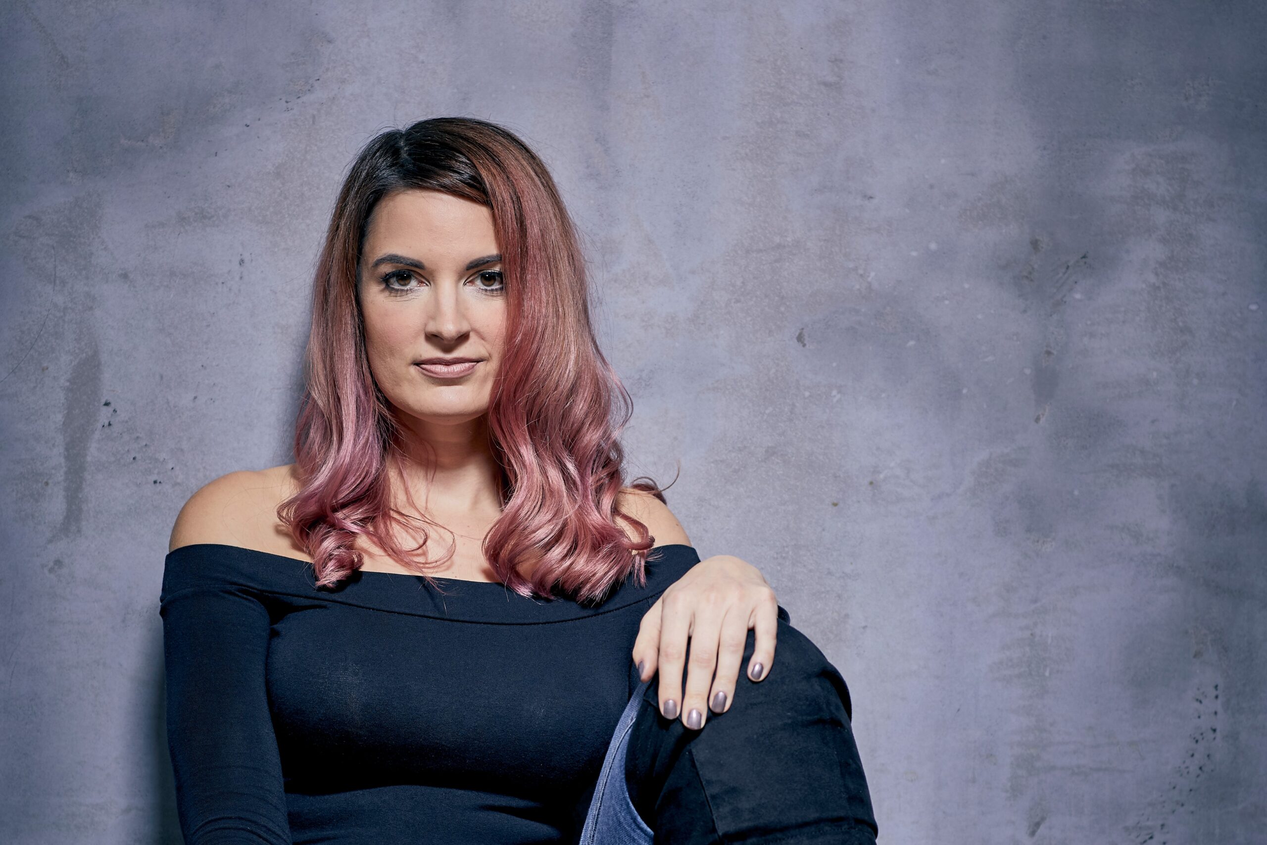 Jazz singer Joanna Hudson sits against a grey background, with pink shoulder-length hair