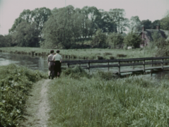 A couple walk alongside a river towards a wooden bridge in a photo of the countryside from 1956