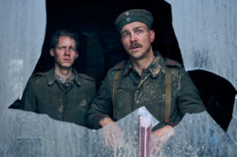 A pair of actors dressed in World War 1 era German soldier's uniforms stare out of a broken window